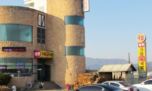 예전 외관 — 석재 외벽 건물, 장작더미, 노란 기둥 간판